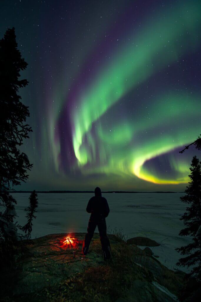 Man standing be campfire below northern lights